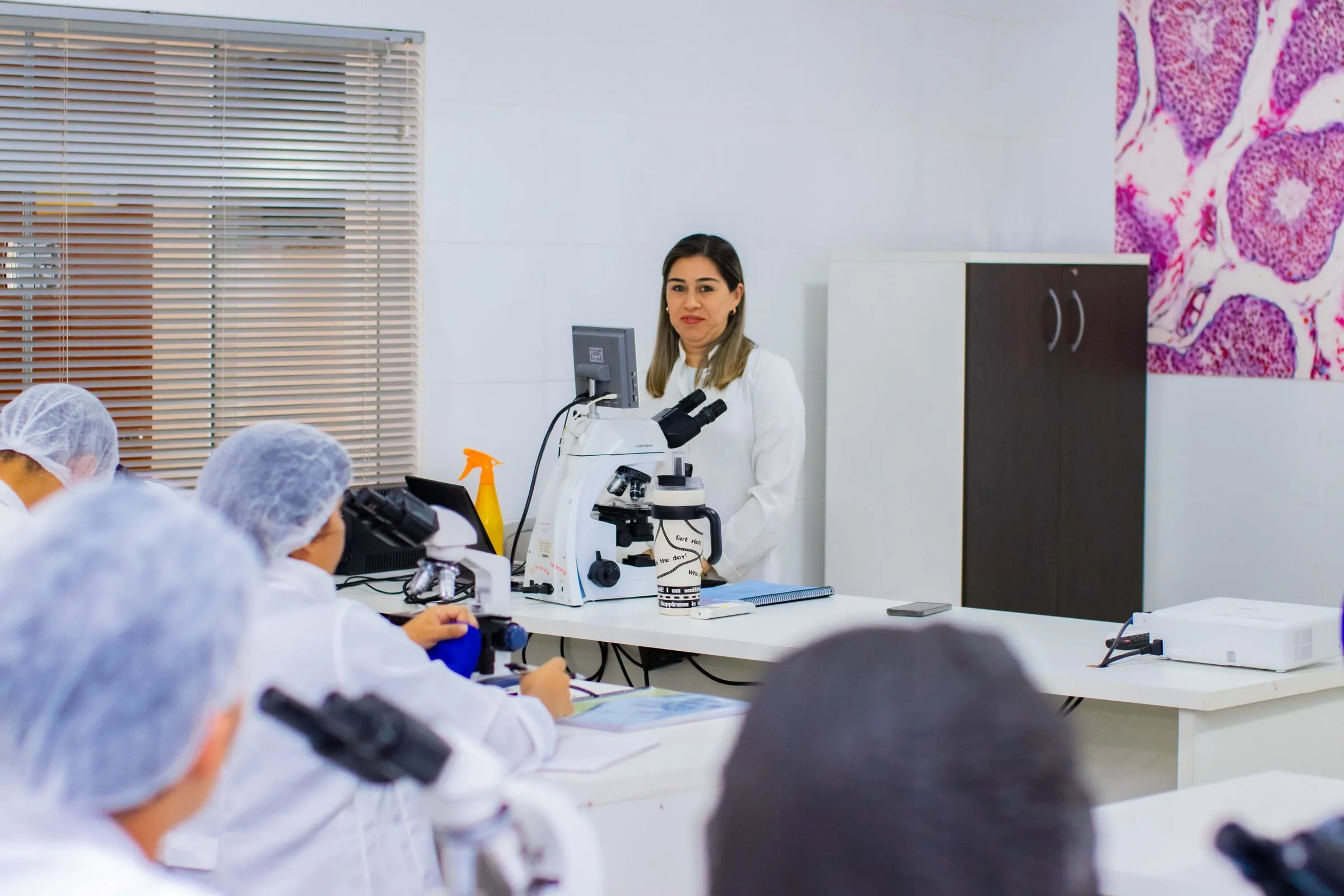 Professora ao microscópio com a imagem ampliada em um monitor, e estudantes sentados em primeiro plano